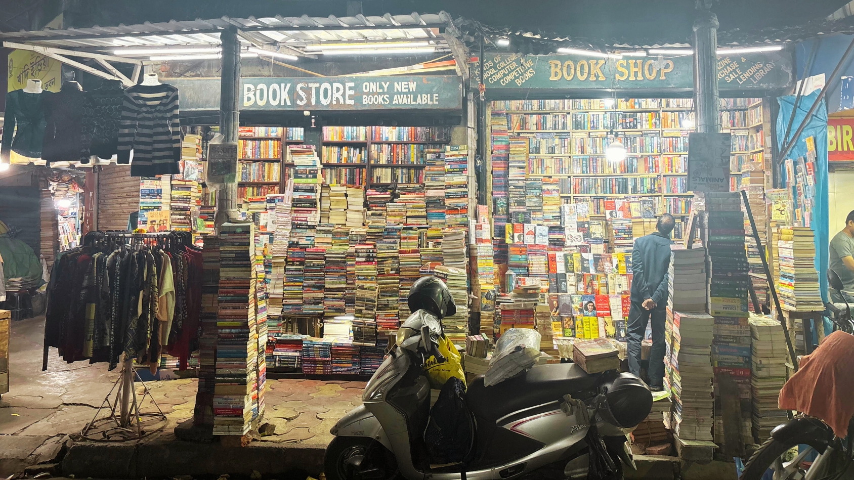 <p>Towering stacks of books at two well-lit street-side bookstores.</p>