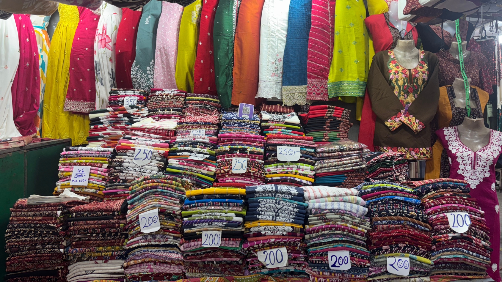 <p>Towers of printed kurtis are stacked with their prices scribbled by hand. </p>